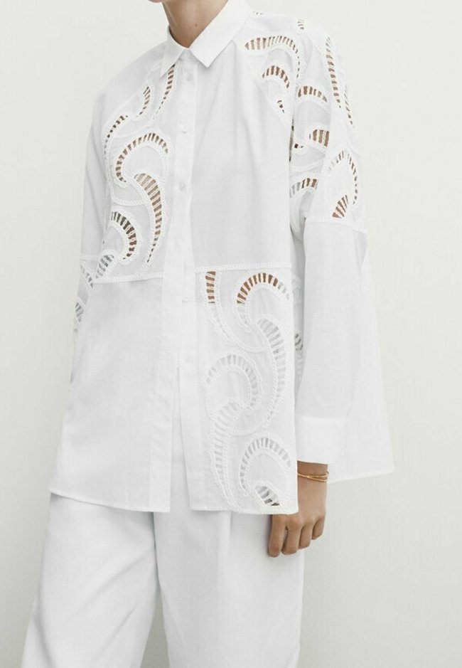 Blouse blanche présentant des motifs découpés complexes et une patte de boutonnage sur le devant. Manches longues avec une coupe ample. Fabriquée en tissu léger.