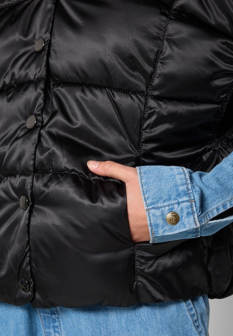 Gilet imbottito nero in tessuto lucido, con bottoni a pressione neri e una mano in una camicia di denim azzurro chiaro con accessori dorati.