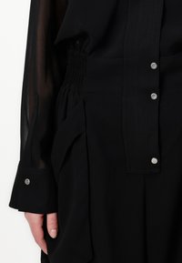 Crna odjeća s smockiranim strukom, prednjim zatvaranjem na gumbe i prozirnim dugim rukavima. Glatka tekstura s metalnim dodacima i strukturiranim dizajnom.