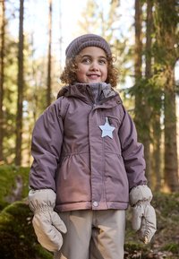 Lilla polstret jakke med hette, grå krage og en stjernebadge. Beige votter og bukser fullfører vinterantrekket, satt i en skogsbakgrunn.