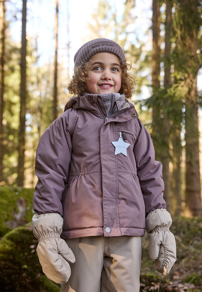 Lilla polstret jakke med hette, grå krage og en stjernebadge. Beige votter og bukser fullfører vinterantrekket, satt i en skogsbakgrunn.