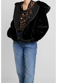 Zwarte nepbont hoodie met een ritssluiting aan de voorkant, gecombineerd met een bruine blouse met luipaardprint en een blauwe spijkerbroek. Zachte textuur met een comfortabele pasvorm.