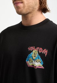 Crni pamukasta majica s šarenim izvezenim dizajnom skeletnog lika koji čita knjigu, s natpisom "VOLCOM" u crvenoj boji iznad grafike.