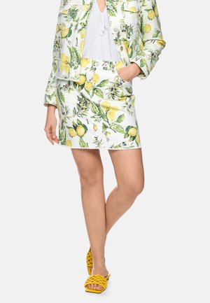 Frau trägt weißen Rock und Jacke mit gelbem Zitronen- und grünem Blattmuster, kombiniert mit gelben offenen Sandalen und weißer Bluse.