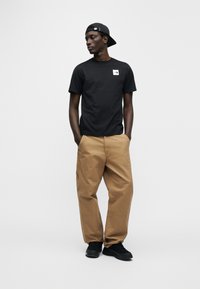 Μαύρο βαμβακερό μπλου ζάκι με λευκό λογότυπο στο στήθος, συνδυασμένο με μπεζ cargo παντελόνι και μαύρα αθλητικά παπούτσια. Άτομο φορά μαύρο καπέλο.