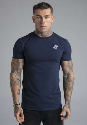 Mann mit tätowierten Armen und Hals trägt ein tailliertes, marineblaues T-Shirt mit weißem Logo auf der Brust, steht vor einem grauen Hintergrund.