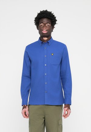 Chemise bleu à manches longues en coton avec col boutonné, poche frontale et logo jaune sur le côté supérieur gauche.