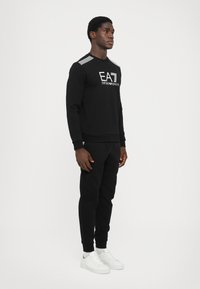 Black sweatshirt with white logo and striped details on the shoulders, paired with matching black joggers and white sneakers.