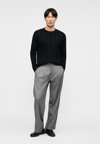 Homme aux cheveux noirs portant un pull texturé noir, un pantalon gris plissé et des chaussures noires, debout devant un fond clair uni.