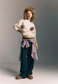 Žena v klobúku s kožúškom, skrátenom krémovom svetre, širokých džínoch, pruhovanej košeli viazanej v páse a hnedých šľapkách pózuje pred jednoduchým pozadím.