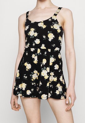 Femme portant une robe noire sans manches à fleurs jaunes et blanches avec jupe plissée, montrée des épaules jusqu'à mi-cuisse.