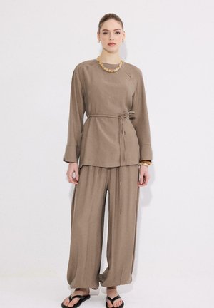 Νέα γυναίκα στέκεται, φοράει φαρδύ τοπ με μακριά μανίκια σε χρώμα ταουπέ με ζώνη στη μέση, παντελόνια με φαρδιά μπατζάκια, κολιέ με χάντρες, βραχιόλια και μαύρα σανδάλια.