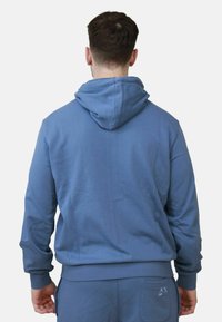 Felpa blu realizzata in morbido tessuto, con cappuccio regolabile, polsini a coste e tasca a marsupio. La vista posteriore mostra un design senza cuciture e dettagli raffinati.