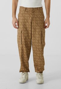 Pantaloni cargo maro cu un model repetitiv "KARL", dotati cu buzunare laterale și manșete elastice la glezne, asociați cu adidași albi.