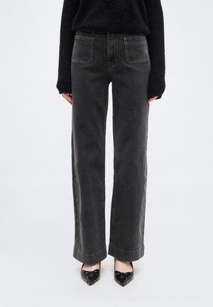 Femeie purtând blugi gri închis cu talie înaltă și picioare largi, cu buzunare aplicate în față, pulover negru pufos și balerini negri cu vârf ascuțit și funde.