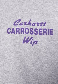 Grå stof med lilla tekst, der læser "Carhartt CARROSSERIE Wip" centreret i tre linjer.