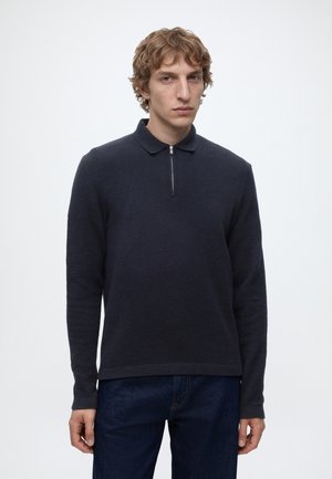 Jeune homme aux cheveux courts et bouclés portant un polo à manches longues bleu marine foncé avec une fermeture éclair à l'avant et un jean bleu foncé sur fond uni.