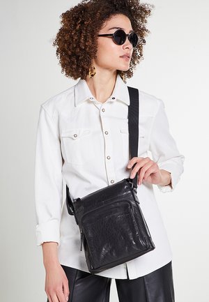 Čierna kožená crossbody taška s textúrovaným povrchom, prednou zipsovou kapsou a nastaviteľným popruhom, držiacou modelom v bielej košeli.