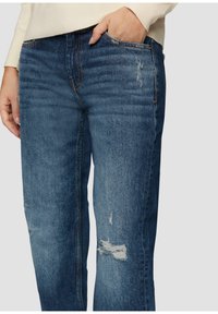 Niebieskie dżinsy o luźnym fasonie, z efektami przetarcia i kontrastowym przeszyciem. Kieszenie widoczne na wysokości talii.