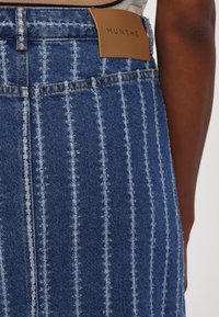 Blauwe denim rok met verticale witte strepen, voorzien van een bruine leren label met "MUNTHE" op de tailleband, en rafelige randen voor textuur.