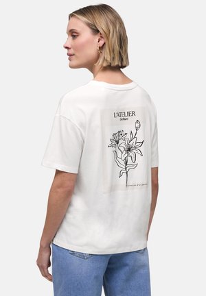 Blondynka w białej koszulce z czarną kwiatową ilustracją i napisem „L'ATELIER De Fleurs” na plecach, w połączeniu z jasnoniebieskimi dżinsami.