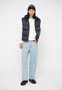 Zils pūkains veste virs baltas kapuces, gaiši zilas platas bikses un melni sporta apavi. Aksesuāri ar rakstainu adītu beanie.