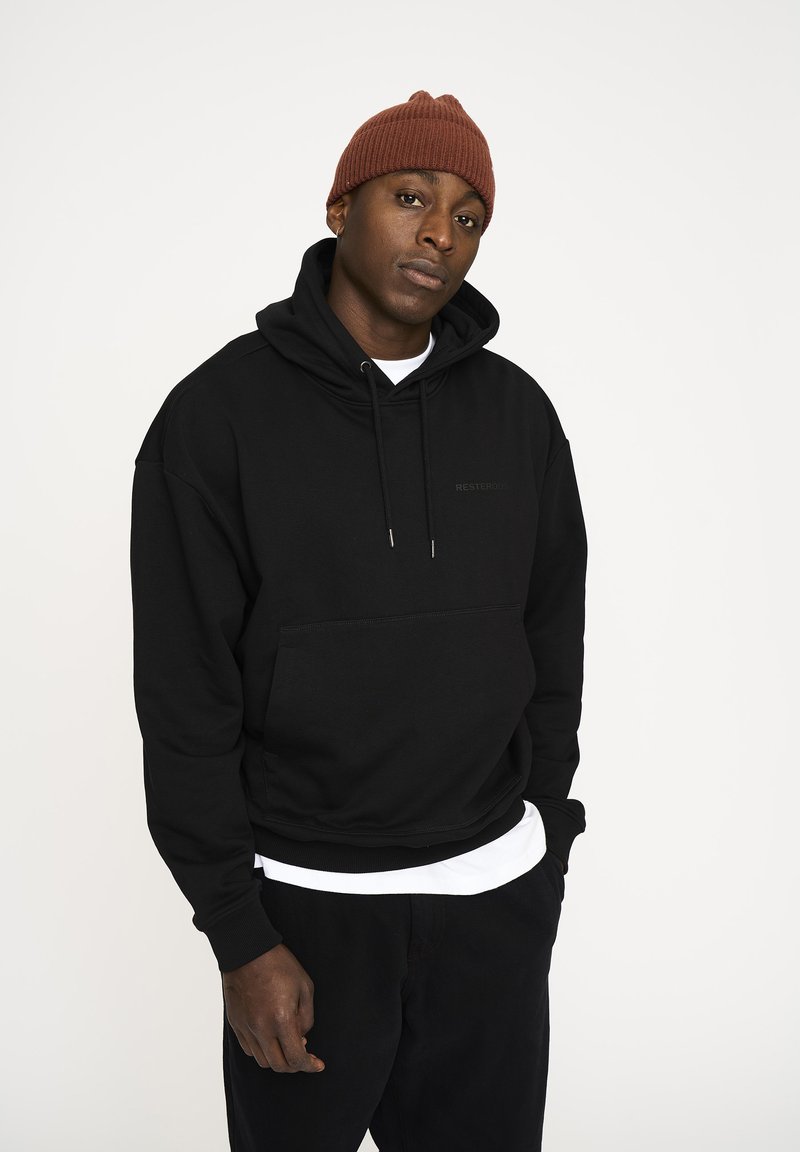 Ung mand iført en brun strikket hue, sort hættetrøje, hvid skjorte under, og sorte bukser, stående med den ene hånd i lommen mod en hvid baggrund.