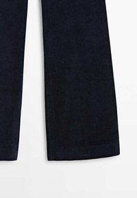 Gros plan sur les jambes d'un pantalon en velours côtelé bleu foncé montrant une texture côtelée verticale et des ourlets droits sur fond blanc.