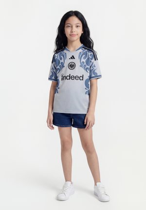 Junges Mädchen mit langen dunklen Haaren, das ein grau-blau gemustertes Sporttrikot, dunkle Shorts und weiße Turnschuhe trägt, steht vor einem einfarbigen Hintergrund.