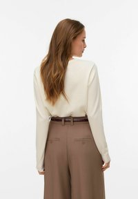 Haut à manches longues couleur crème associé à un pantalon beige taille haute avec une ceinture texturée et deux poches arrière avec détail bouton.