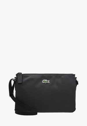 Czarna torba Lacoste typu crossbody z teksturowaną powierzchnią, małym zielonym logo krokodyla, regulowanym paskiem oraz zamkiem błyskawicznym na górze.