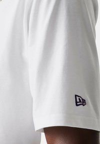 T-shirt bianco a maniche corte realizzato in cotone. Presenta un logo ricamato viola sul polsino. Texture liscia e vestibilità standard.