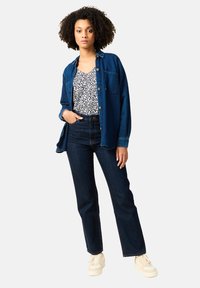Camicia di jeans blu scuro sopra un top a fiori, abbinata a jeans neri a vita alta e sneaker beige. Maniche lunghe e vestibilità comoda.