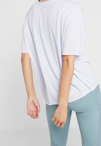 Camiseta blanca de corte relajado y mangas cortas, con un pequeño logo de Nike en la parte superior de la espalda, combinada con leggings en color teal.