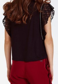 Top nero a maniche corte con accenti in pizzo sulle spalle, dal design pulito e una texture liscia, abbinato a pantaloni rossi.