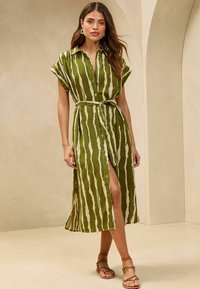 Robe chemise rayée verte avec une taille ceinturée, des manches courtes, un bouton à l'avant, et une fente sur le côté, associée à des sandales brunes à lanières.
