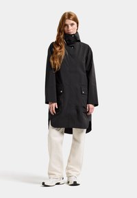 Svart vattentät parka med hög krage, framknäppningar och sidofickor, bärs över ljusa byxor och sneakers.