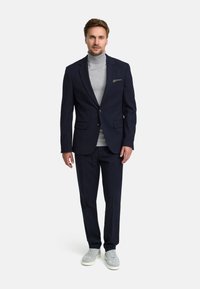 Marine blazer met een enkelvoudige sluiting en bijpassende broek, gecombineerd met een lichtgrijze turtleneck en grijze sneakers. Glad materiaal.