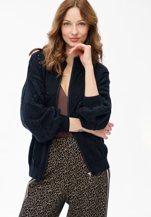 Młoda kobieta o falujących brązowych włosach, ubrana w czarny dziergany kardigan, brązową bluzkę i spodnie w panterkę, pozuje z dłonią przy brodzie na białym tle.