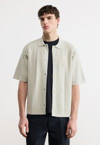 Νέος άντρας που φοράει ένα μπεζ πουκάμισο με κοντά μανίκια και κουμπιά πάνω από ένα μαύρο τοπ, στέκεται μπροστά σε ένα απλό λευκό φόντο.