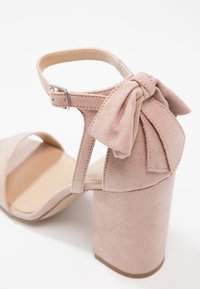 Sandale à talon bloc en suède rose clair avec bride à la cheville, boucle argentée et nœud décoratif en tissu à l'arrière.