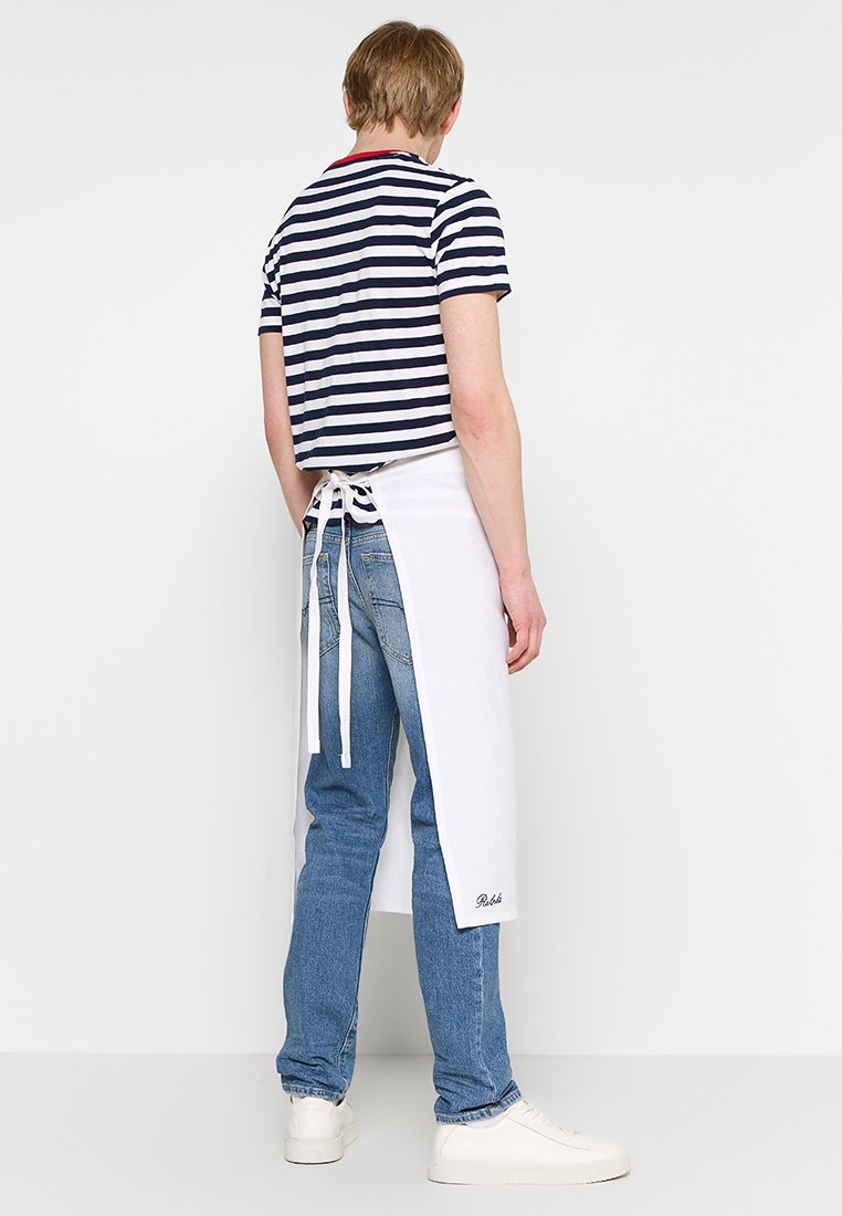 Grembiule bianco con lacci, indossato sopra jeans blu e una maglietta a righe navy. Presenta un dettaglio con logo. Lo sfondo è neutro. Il modello è girato di spalle.