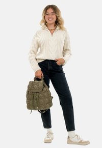 Femme souriante, portant un pull en tricot crème avec motifs en relief, un jean noir, des baskets blanches, et tenant un sac à dos matelassé olive vert par les poignées.