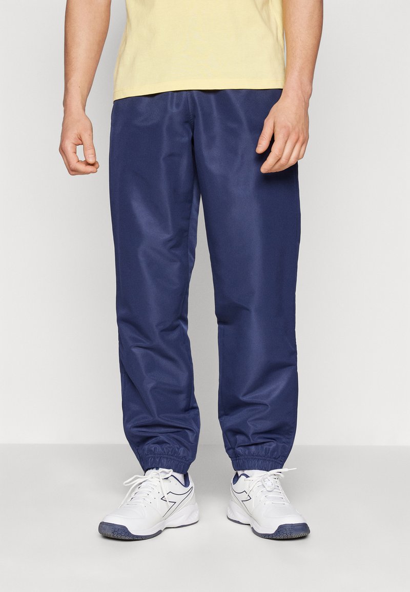 Marineblå joggebukser med elastisk midje og mansjetter, laget av glatt, lett materiale. Kombinert med hvite joggesko.