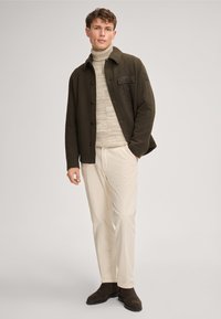 Wełniana kurtka w oliwkowej zieleni z zapięciem na guziki, kieszenią na piersi, kremowy dziergany golf, jasne spodnie oraz brązowe zamszowe botki za kostkę.