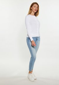 Witte lange mouwen cardigan met knopen, ribbelzoom, gecombineerd met lichtblauwe skinny jeans en witte sneakers, tegen een effen achtergrond.
