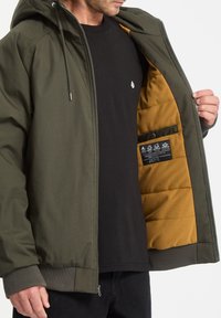 Grøn isoleret jakke med lynlås, hætte og elastiske manchetter. Foret med gult materiale; har et plejelabel i lommen indeni. Sort t-shirt under.