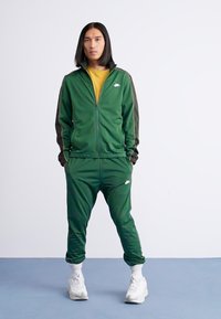 Un survêtement vert en matériau lisse comprend une veste zippée avec des accents rayés et un pantalon assorti, associé à des baskets blanches.