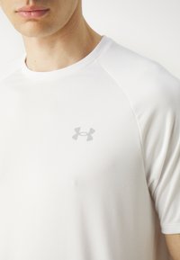 Camiseta deportiva blanca con mangas raglán cortas, textura suave, con un sutil logo gris de Under Armour en el pecho.