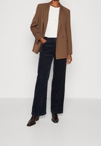 Hnědý saka na míru přes bílé přiléhavé tričko, spárované s tmavě modrými manšestrovými kalhotami širokého nohavicového střihu a tmavými kotníkovými botami na blokovém podpatku.